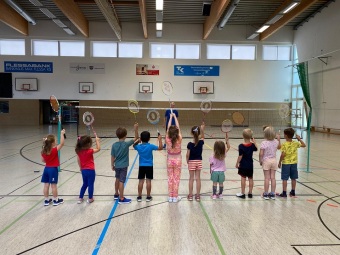 Kinder stehen in einer Sporthalle und halten Badmintonschläger hoch.